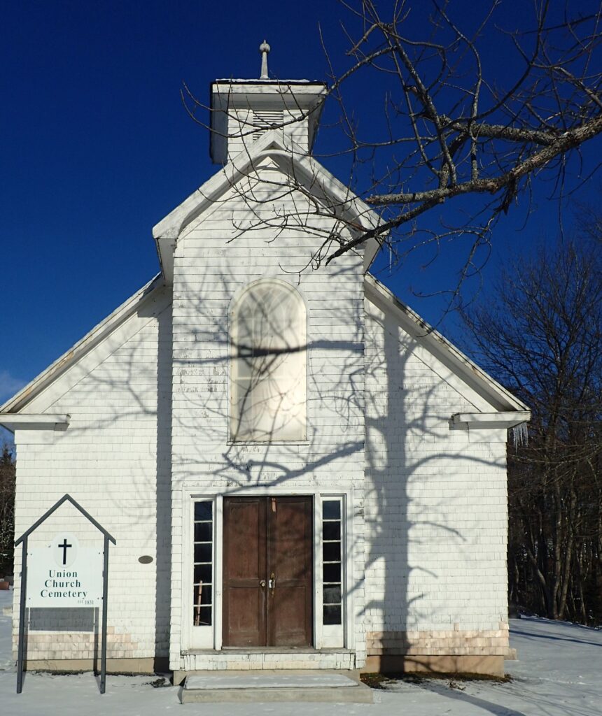 A white church with a small steeple and the shadow of a leafless tree cast over the front.