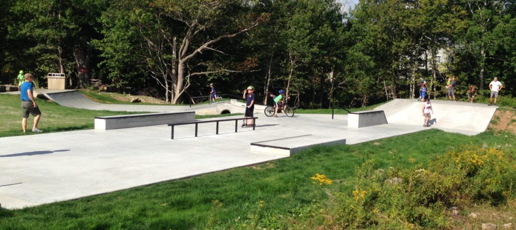 People using a concrete skate park surrounded by green grass and trees.