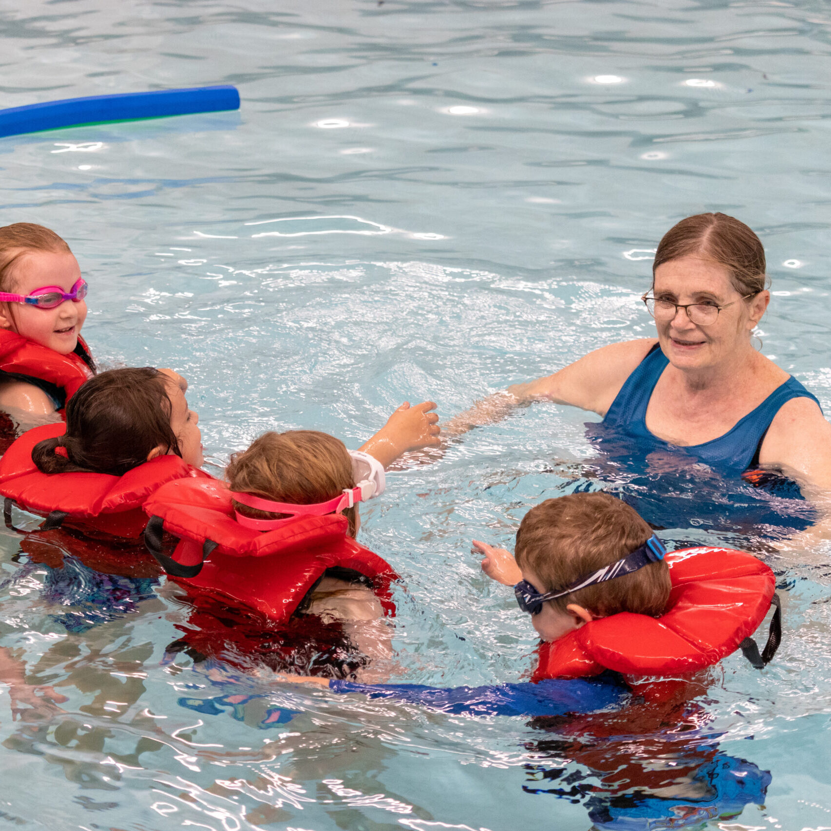 A lifeguard leads a group of young children wearing lifejackets in a swimming lesson