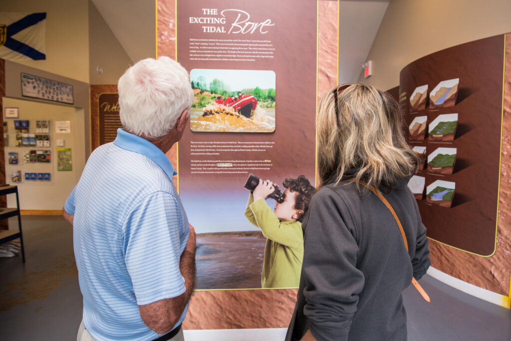 Two people with their backs to the camera stand reading an information display inside the Fundy Tidal Interpretive Centre.