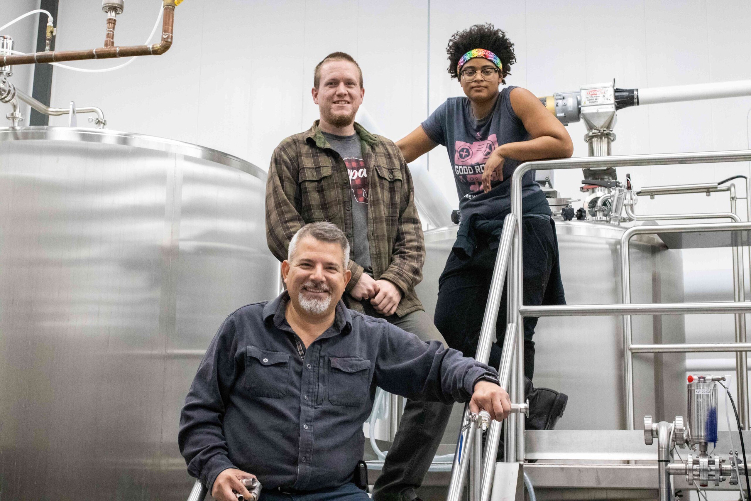 Three people stand smiling on steps next to stainless steel brewing equipment