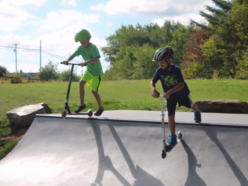 Two young children on scooters using a skate park ramp
