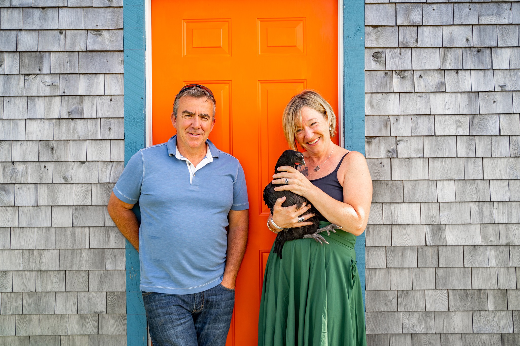 Two people stand in front of a bright orange door. The person on the right is holding a black chicken.