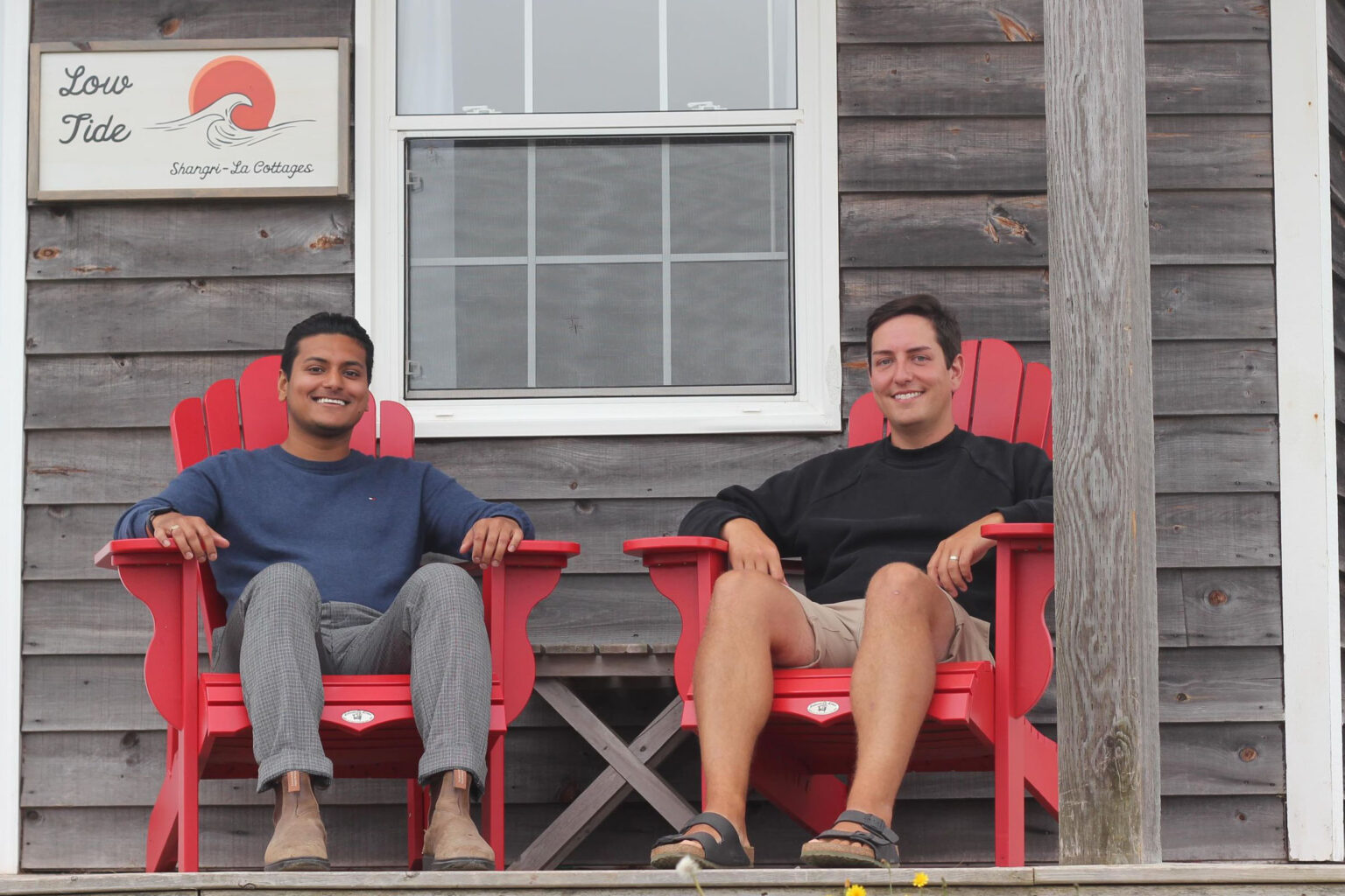 Two smiling people sit in red Adirondack chairs on the deck of a cottage.