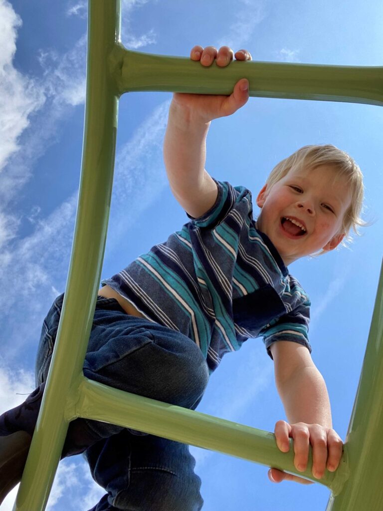 A blonde child smiles down at the camera while climbing on a green ladder