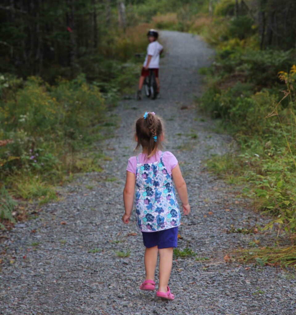 A young girl walking on a gravel path away from the camera. A young boy on a bike is ahead of her looking back.